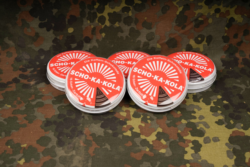 5 Zartbitter Schokakola Dosen aufgestapelt vor einem Flecktarn Hintergrund